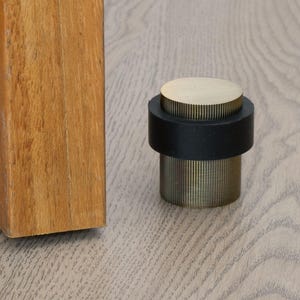 May include: A brass and black door stop with a cylindrical design. The top is a flat, circular brass piece, and the base is a ribbed, cylindrical brass section. A black rubber ring sits between the top and base. The door stop is on a light wood floor.