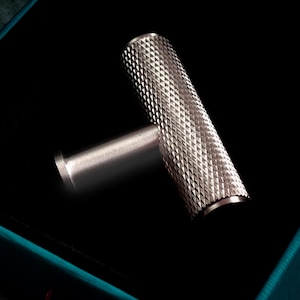 Puede incluir: Un tirador de armario plateado con un mango texturizado con patrón de diamantes. El tirador en forma de T está sobre un fondo negro dentro de una caja verde azulado. El tirador de metal tiene un extremo curvo.