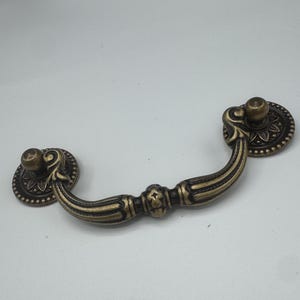 May include: Antique brass drawer pull with decorative floral detailing. The pull has a curved handle and two circular backplates with raised floral patterns. The hardware is designed for furniture and cabinets.