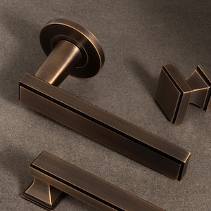 Puede incluir: Un juego de herrajes para puertas en tono bronce, que incluye una manija de palanca, un pomo cuadrado y un tirador rectangular. Cada pieza presenta una línea de acento oscuro. Para puertas y armarios.