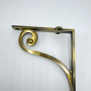 Puede incluir: Un soporte para estante en forma de L hecho de metal con tono de latón antiguo. El soporte presenta un brazo de apoyo curvo en forma de voluta con un detalle circular central. Incluye orificios de montaje preperforados en las secciones.