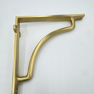 Puede incluir: Un soporte para estante de metal en color dorado con un marco en forma de L y un refuerzo curvo. El soporte cuenta con superficies de montaje planas con orificios para tornillos preperforados en los extremos superior y lateral, con acabado mate.