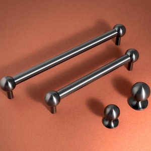 May include: A collection of brushed nickel cabinet hardware, featuring two bar pulls and two round knobs. The pulls have a cylindrical design with spherical ends. The knobs have a similar spherical design. The hardware is displayed on a copper-coloured surface.