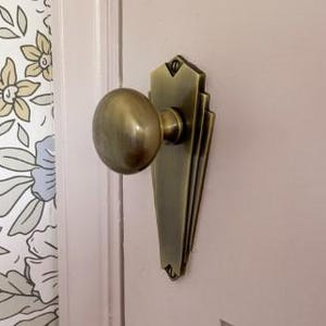Peut inclure: Un bouton de porte et une plaque arrière en laiton sont montés sur une porte rose clair. Le bouton de porte est rond et la plaque arrière a un design géométrique. Papier peint floral aux couleurs pastel visible à gauche.