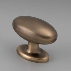 Może przedstawiać: Zestaw mosiężnych okuć do szafek, w tym długi uchwyt i pasujący owalny gałek. Okucia mają gładkie, metaliczne wykończenie i są przeznaczone do mebli lub szafek.