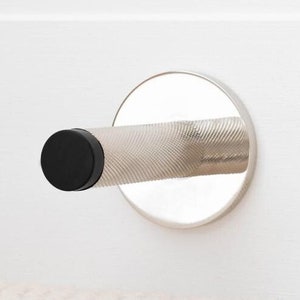 May include: A chrome door stop with a textured handle and a black rubber end, mounted on a white door. The door stop has a circular base and is designed to prevent a door from slamming shut.