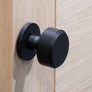 May include: A black door knob with a round design. The knob is mounted on a light wood door.