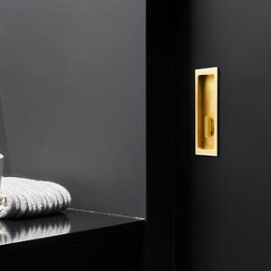 Puede incluir: Interior de baño moderno con un lavabo blanco de forma ovalada, una encimera negra y un mueble negro. Un tirador de puerta empotrado de color dorado es visible en una puerta negra. Una toalla blanca está sobre la encimera.