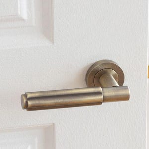May include: A bronze-colored door handle with a curved design. The handle is attached to a white door, with a simple, elegant aesthetic. The handle's finish has a slightly aged appearance, adding a touch of vintage charm to the door.