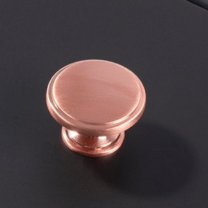 May include: Two copper cabinet knobs with a round, slightly raised top and a small, round base. The knobs are on a black surface.