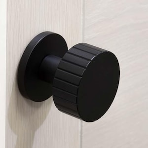 May include: A black door knob with a round, ridged design. The knob is mounted on a white door.
