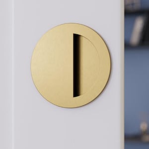 May include: A circular, gold-coloured door pull with a vertical slot for gripping. The hardware is mounted on a white door, with a glimpse of a blue wall and shelving in the background. The door pull has a minimalist design.