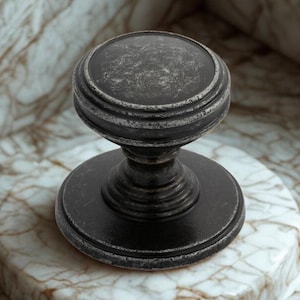 May include: A dark, antique-style cabinet knob with a round top and base. The knob is set on a marble surface with brown and white veining. The knob's finish has a distressed appearance, adding to its vintage charm.