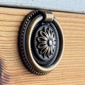 Könnte beinhalten: Nahaufnahme einer Holzkiste mit einem Messingringzug. Der Ringzug hat ein dekoratives Blumenmuster.