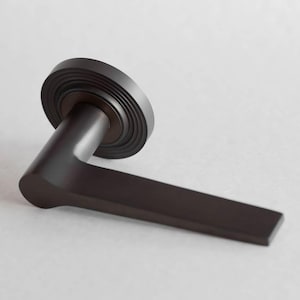 Puede incluir: Una manija de puerta en color negro mate con una palanca horizontal plana y una placa de montaje circular con detalles de anillos concéntricos. El herraje tiene un acabado sólido y liso con un diseño geométrico limpio.