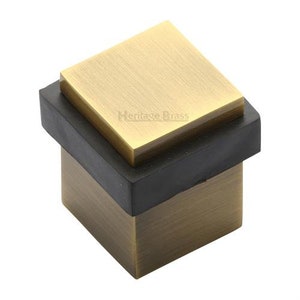 May include: A square doorstop with a brushed brass finish and a black rubber base. The top of the doorstop is a separate square piece of brass. The doorstop is labeled "Heritage Brass".
