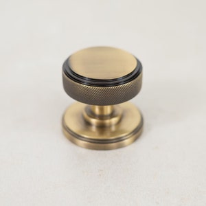 Może przedstawiać: Okrągły, mosiężny klamka do drzwi z czarnym pierścieniem akcentującym i fakturowanym wzorem. Klamka ma polerowane wykończenie.