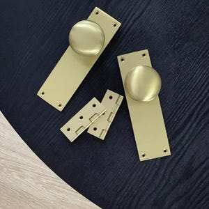 Peut inclure: Un ensemble de quincaillerie de porte en laiton, comprenant deux plaques arri&egrave;re rectangulaires avec des boutons ronds et deux petites charni&egrave;res. La quincaillerie a une finition mate et est dispos&eacute;e sur une surface sombre.