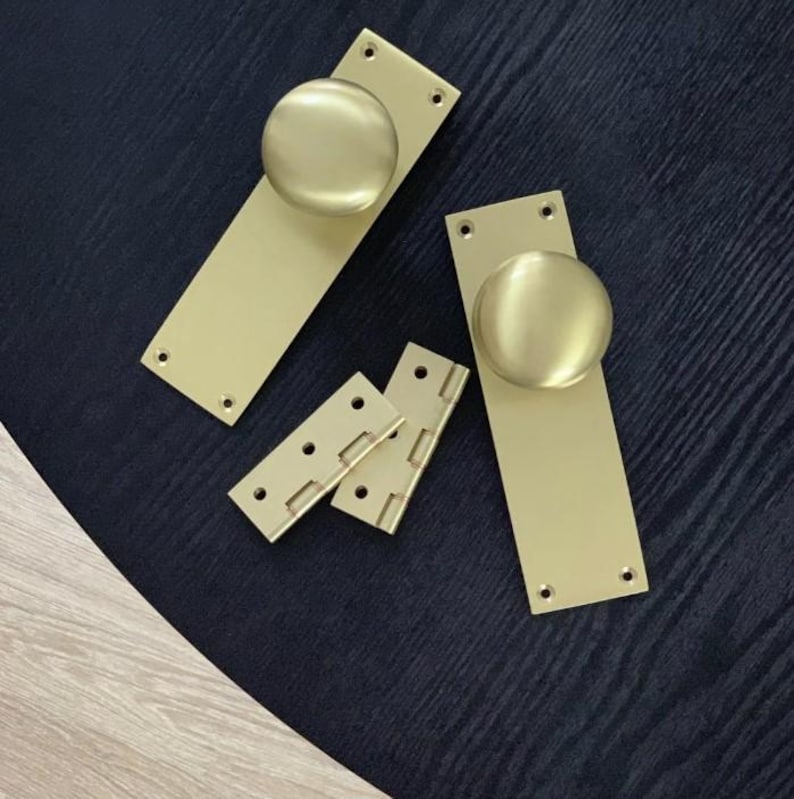 Peut inclure: Un ensemble de quincaillerie de porte en laiton, comprenant deux plaques arri&egrave;re rectangulaires avec des boutons ronds et deux petites charni&egrave;res. La quincaillerie a une finition mate et est dispos&eacute;e sur une surface sombre.