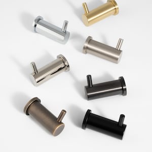 May include: Seven metal wall hooks in different finishes: silver, gold, bronze, and black. Each hook has a cylindrical body with a small, projecting hook. The hooks are displayed on a white surface.