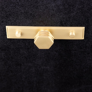 Puede incluir: Un tirador de armario dorado con una placa trasera rectangular y dos pomos redondos. El tirador central tiene forma hexagonal. El herraje es de metal sobre un fondo oscuro.