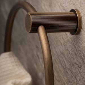 Könnte beinhalten: Ein bronzefarbener Handtuchring mit einer zylindrischen Stange mit horizontalen Rillen. Der Ring ist an einer strukturierten, grauen Wand befestigt. Ein weißes Handtuch ist teilweise sichtbar.