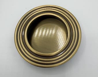 Könnte beinhalten: Ein runder, eingelassener Griff mit gebürsteter Messingoberfläche. Das Beschlagteil verfügt über ein gestuftes, konzentrisches Ringdesign, das eine zentrale konkave Vertiefung umgibt und als Griffmulde für Schiebetüren oder Schränke dient.