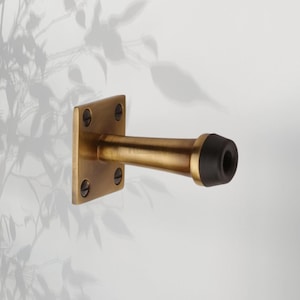 Peut inclure: Butoir de porte en laiton avec une base carrée et un corps cylindrique. La base est fixée avec quatre vis. L'extrémité du butoir de porte est dotée d'un tampon en caoutchouc noir.