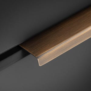 Può includere: Primo piano di una maniglia per cassetto color bronzo con forma rettangolare e un bordo leggermente curvo. La maniglia è posta su una superficie grigio scuro, creando un contrasto di toni. L'hardware è probabilmente per un armadio o un cassetto.
