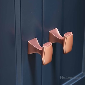 Può includere: Due pomelli per armadio in rame con forma rettangolare e design curvo. I pomelli sono montati su una porta di armadio blu scuro.