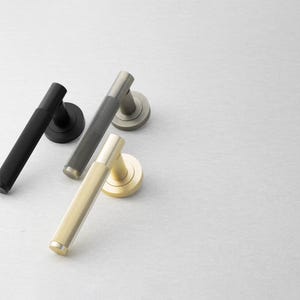 May include: A collection of door handles in various metallic finishes. The handles are black, gray, and gold, each with a textured, cylindrical grip and a circular base. The handles are arranged on a light gray surface.