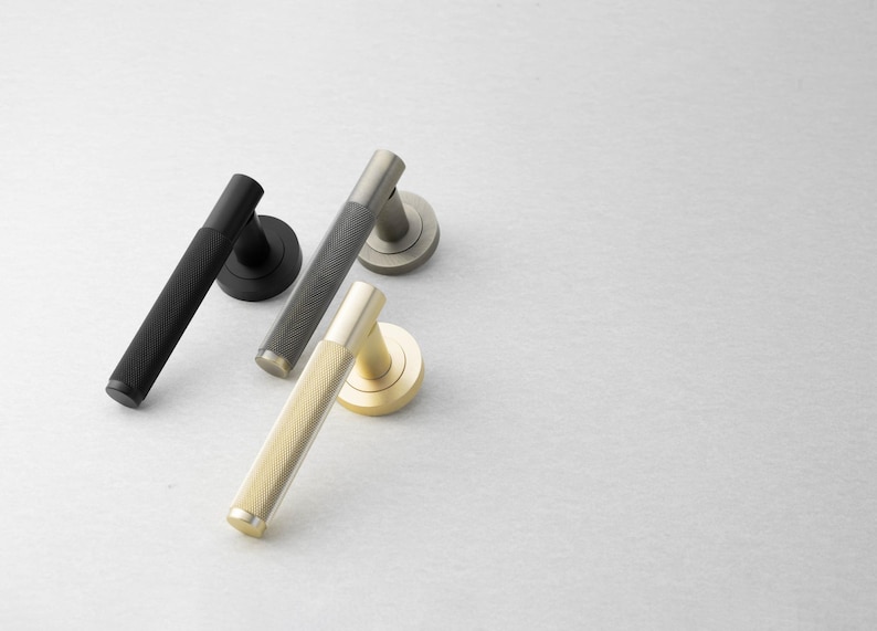May include: A collection of door handles in various metallic finishes. The handles are black, gray, and gold, each with a textured, cylindrical grip and a circular base. The handles are arranged on a light gray surface.
