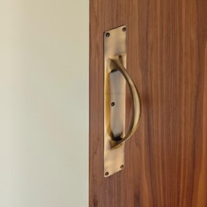 May include: A brass door pull with a curved handle is mounted on a wooden door. The rectangular backplate is secured with four screws. The door is a warm brown color, and the wall to the left is a light beige.