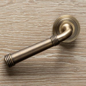 Op de afbeelding: Close-up van een antieke messing deurklink. De klink heeft een gebogen hendelontwerp met een ronde achterplaat. De afwerking is een warme, verouderde messingkleur, met gedetailleerde ribbels.