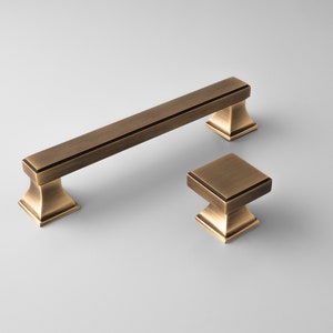 Peut inclure: Deux poignées de porte en laiton antique. L'une est une longue poignée de barre avec une base carrée et l'autre est un bouton carré plus petit avec une base carrée.