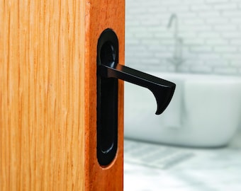 May include: A matte black recessed sliding door handle with a protruding lever mechanism installed into the edge of a light-toned wooden door. The handle features an elongated oval plate and a horizontal handle grip extending outward.