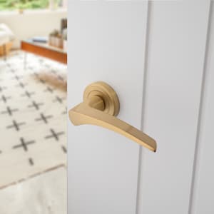 May include: A gold door handle with a curved lever and a round, circular base. The handle is mounted on a white door.