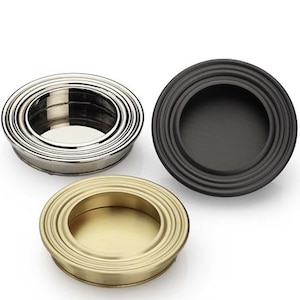 May include: Three circular cabinet pulls in different finishes: polished silver, matte black, and brushed gold. Each pull has a recessed center and concentric rings around the edge. The pulls are designed for easy finger access.