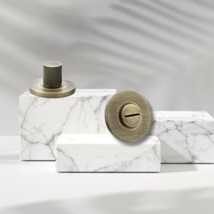 May include: Two antique brass cabinet knobs with a round base and a raised, textured knob. The knobs are displayed on white marble platforms.