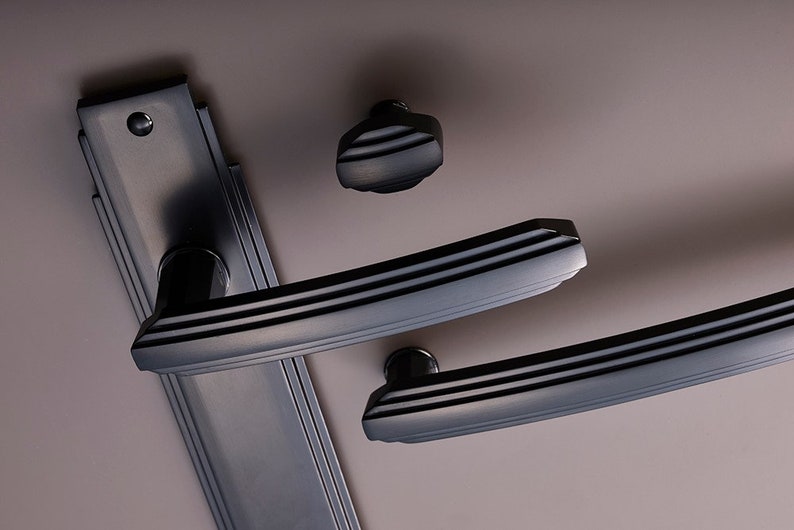 K&ouml;nnte beinhalten: Schwarzes T&uuml;rgriff-Set mit einer rechteckigen Grundplatte und zwei Hebelgriffen. Die Griffe haben einen dekorativen Art-Deco-Stil.