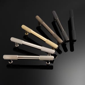 May include: An assortment of cabinet pulls in various metallic finishes, including silver, gold, bronze, and black. Each pull features a textured, cylindrical handle and is mounted on a dark, reflective surface.