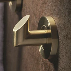May include: A gold-colored metal wall hook with a circular base and an angled hook. The hook is mounted on a textured, brown wall. The design is modern and minimalist, suitable for hanging towels or robes.