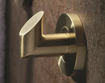May include: A gold-colored metal wall hook with a circular base and an angled hook. The hook is mounted on a textured, brown wall. The design is modern and minimalist, suitable for hanging towels or robes.