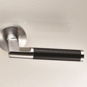 May include: A silver door handle with a black carbon fiber insert. The handle has a round base and a long, rectangular lever.