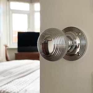 Könnte beinhalten: Nahaufnahme eines polierten verchromten Türknaufs mit rundem, geripptem Design. Der Knauf ist an einer weißen Tür montiert.