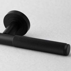 May include: A black door handle with a textured grip and a circular backplate. The handle is made of metal and has a matte finish. The design is modern and minimalist.