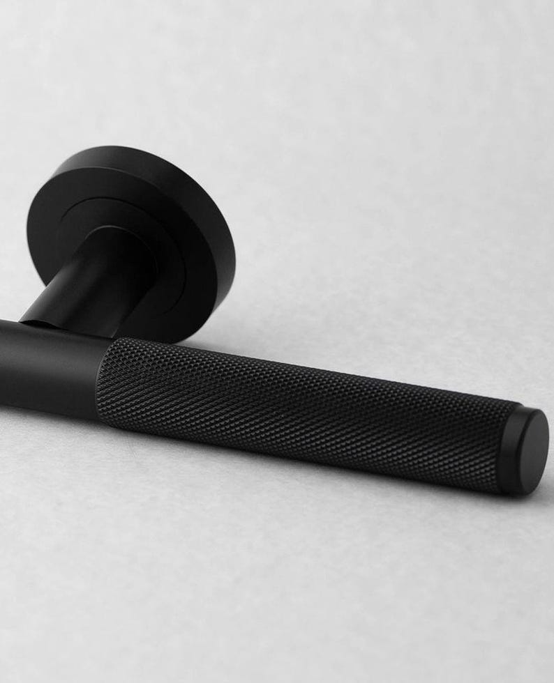 May include: A black door handle with a textured grip and a circular backplate. The handle is made of metal and has a matte finish. The design is modern and minimalist.