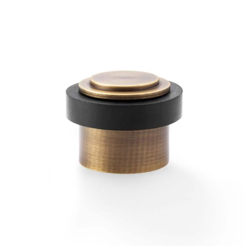 May include: A brass doorstop with a black rubber base. The doorstop has a circular top with a raised ring around the edge.