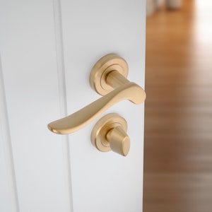 May include: A gold door handle with a curved lever and a round knob. The handle is mounted on a white door.