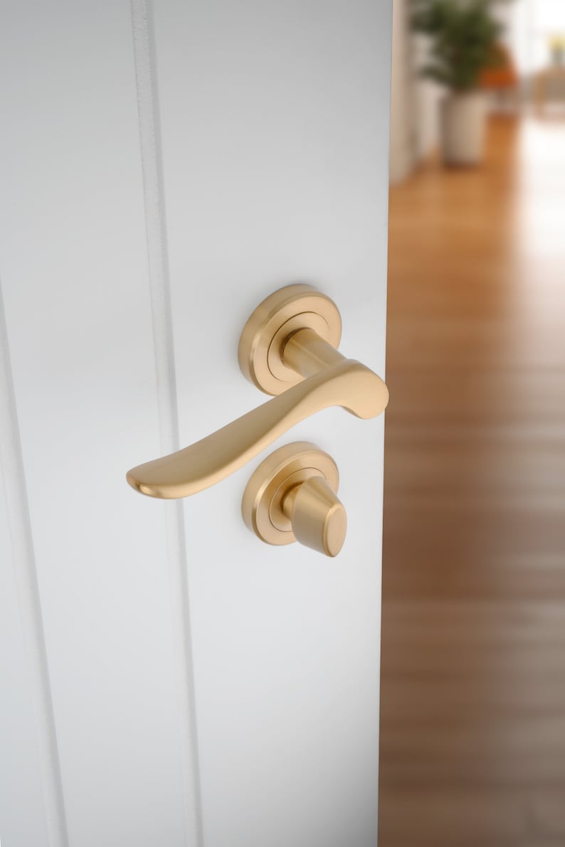 May include: A gold door handle with a curved lever and a round knob. The handle is mounted on a white door.